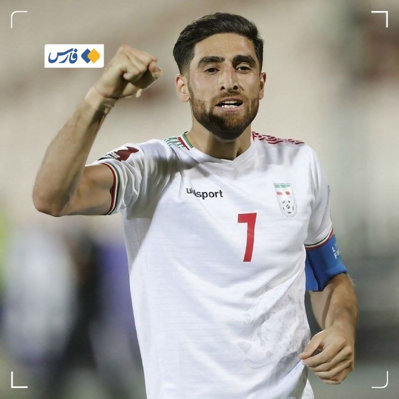 A bearded soccer player in a white jersey with the number 7, a team crest, and a blue captain's armband raises his fist in celebration.