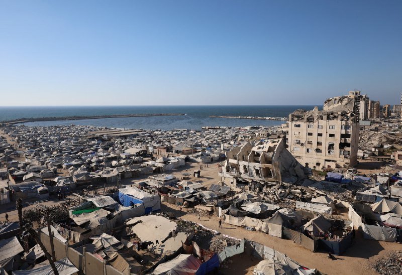 A sprawling encampment of makeshift tents stretches across a coastal area beside heavily damaged multi-story buildings and rubble, with the sea visible in the background.