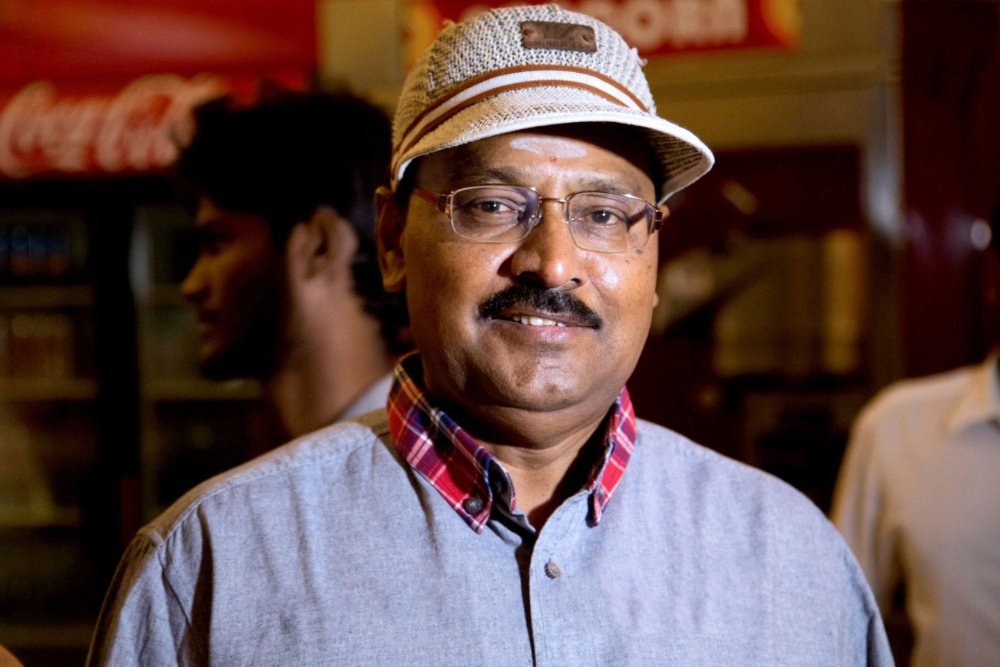 A smiling man wearing glasses, a gray fedora hat, and a checkered shirt over a blue tunic stands indoors, with a Coca-Cola sign visible in the blurred background.