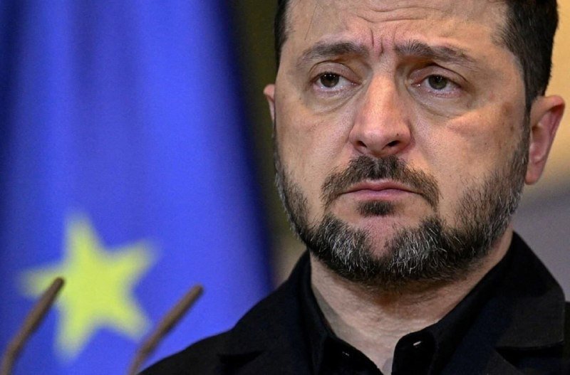 A bearded man in a black shirt looks forward with a serious expression, a blue flag with yellow stars visible behind him.