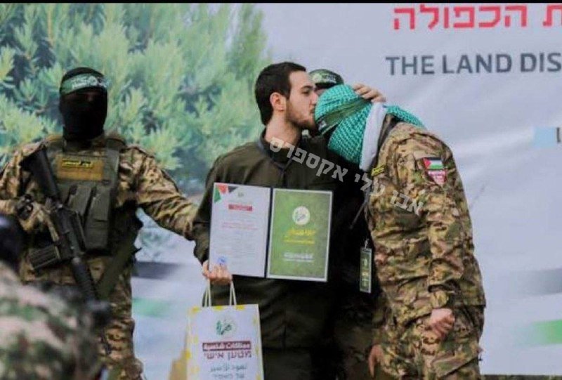 A man kisses the cheek of a masked, keffiyeh-clad figure in camouflage as armed masked fighters stand nearby, with one holding a certificate and gift bag at an outdoor ceremony.