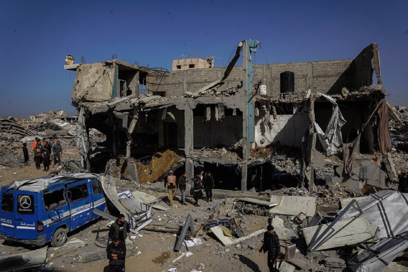 A destroyed multi-story building stands in rubble as people, including some in vests, inspect the wreckage near a blue police van.