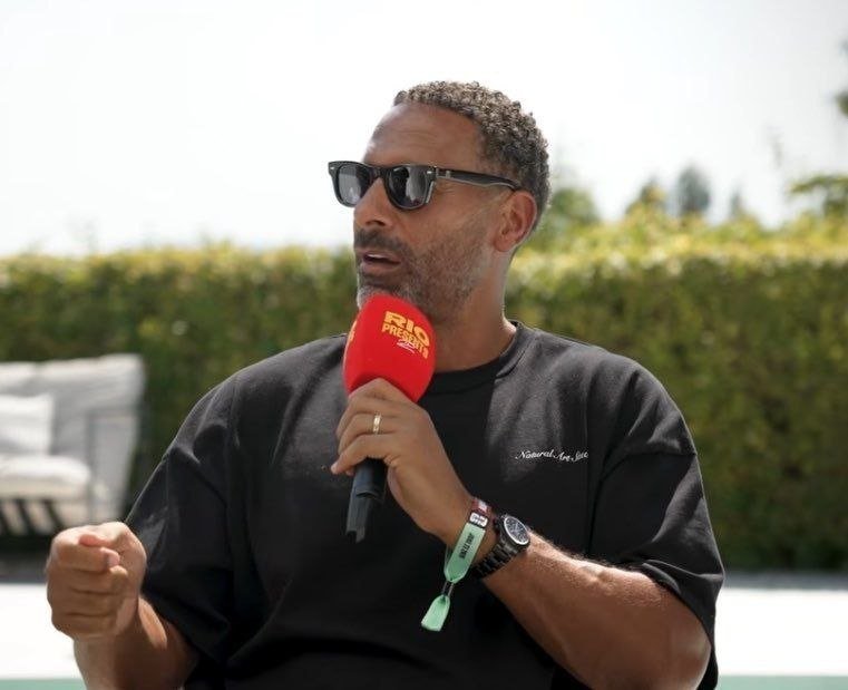 A man wearing sunglasses and a black t-shirt speaks into a red "Rio Ferdinand Presents" microphone outdoors.