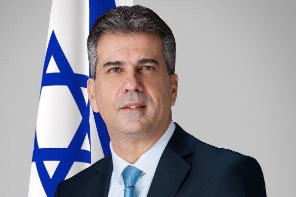 A gray-haired man in a dark suit and light blue tie stands in front of an Israeli flag against a gray background.