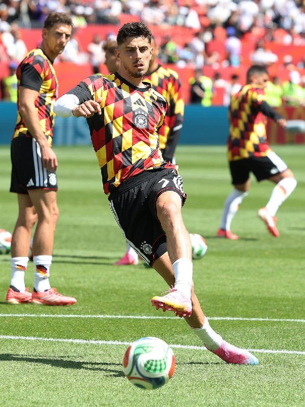 Soccer players in red, yellow, and black diamond-patterned jerseys warm up on a grass field, with one player kicking a ball while others stretch nearby in front of a stadium crowd.