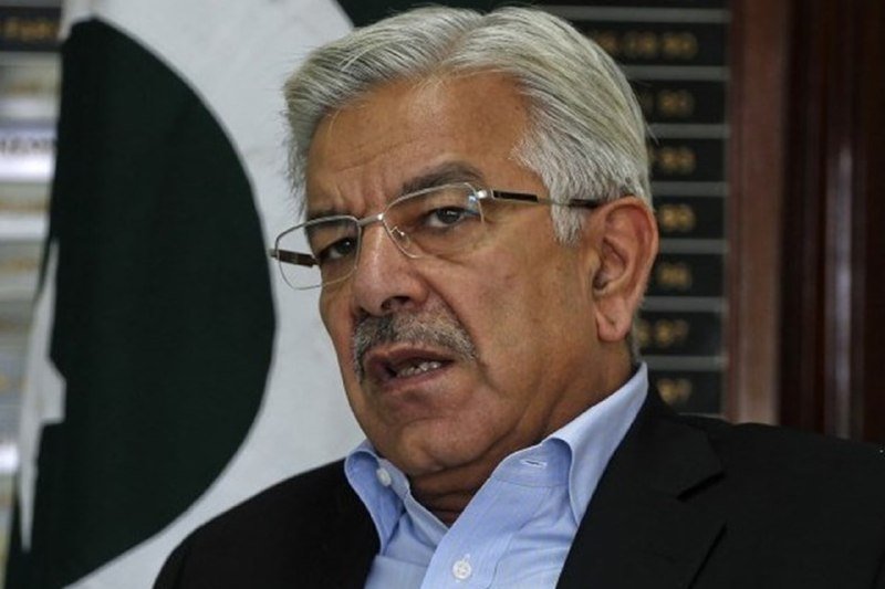 An older man with gray hair and glasses speaks while standing in front of a black-and-white flag and a plaque board.