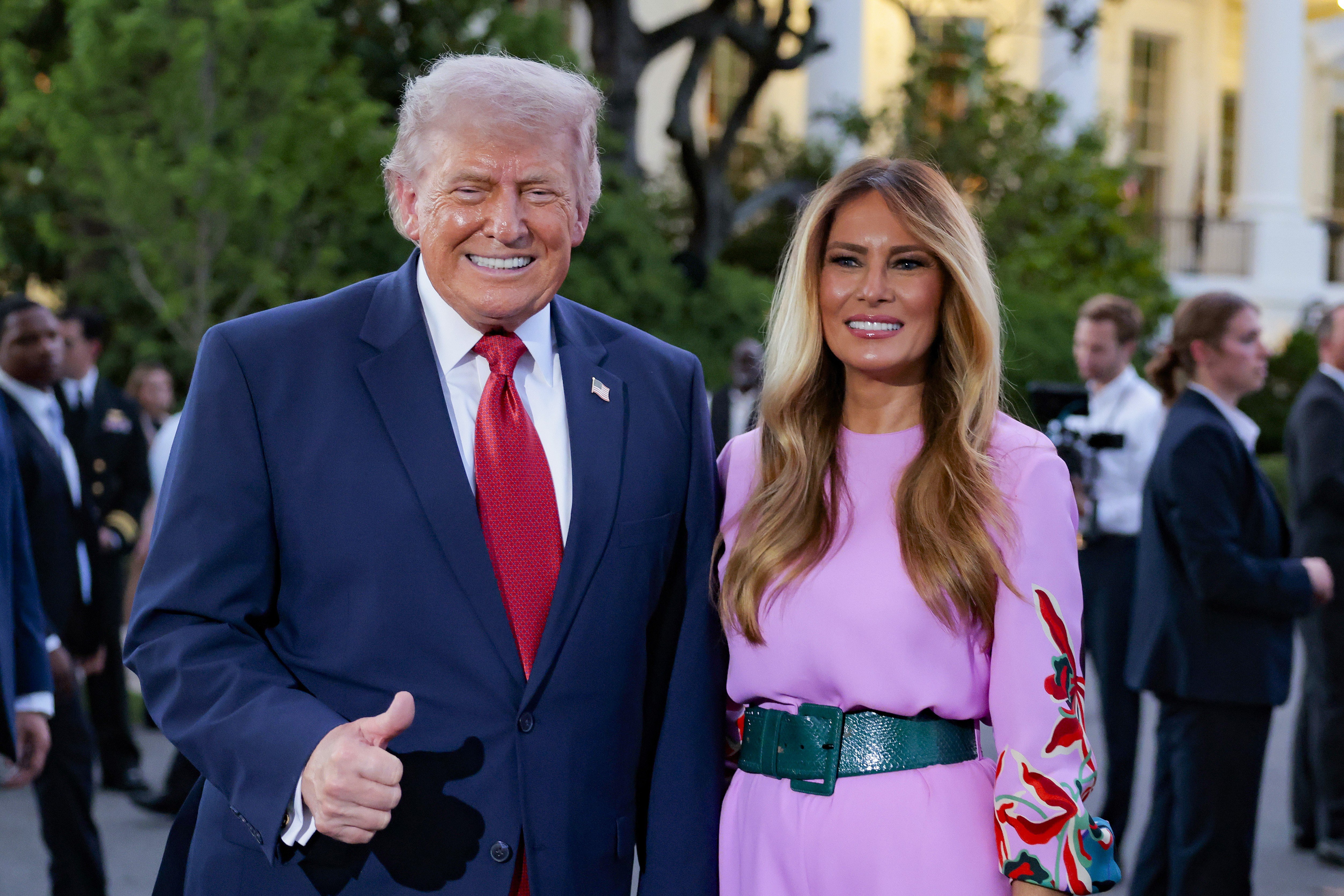 A smiling older man in a navy suit gives a thumbs-up beside a woman in a pink belted dress with floral sleeves.