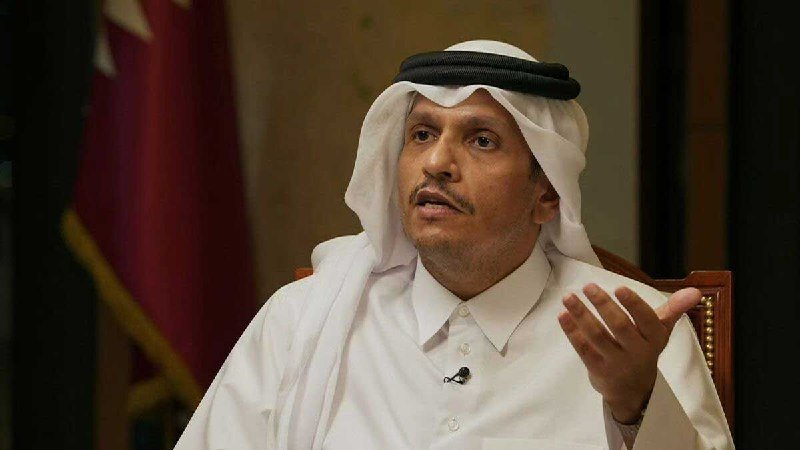 A man wearing a white thobe and ghutra with a black agal gestures with his hand while seated indoors, with a red flag visible behind him.