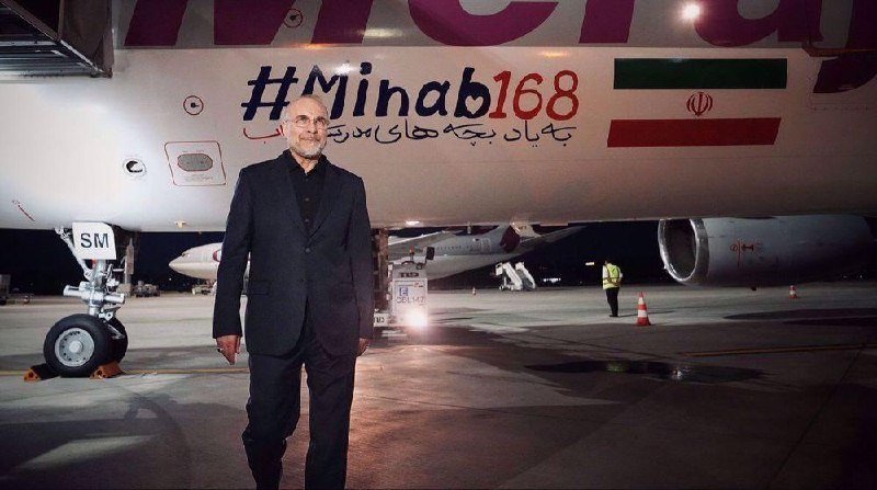 A bearded man in a dark suit stands on an airport tarmac at night in front of an aircraft displaying "#Minab168" and an Iranian flag emblem.