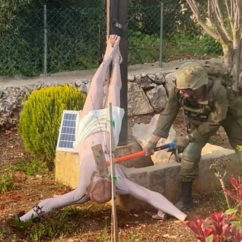 A soldier in combat gear uses a long tool on a mannequin tied upside-down to a wooden post outdoors, with a solar panel visible nearby.