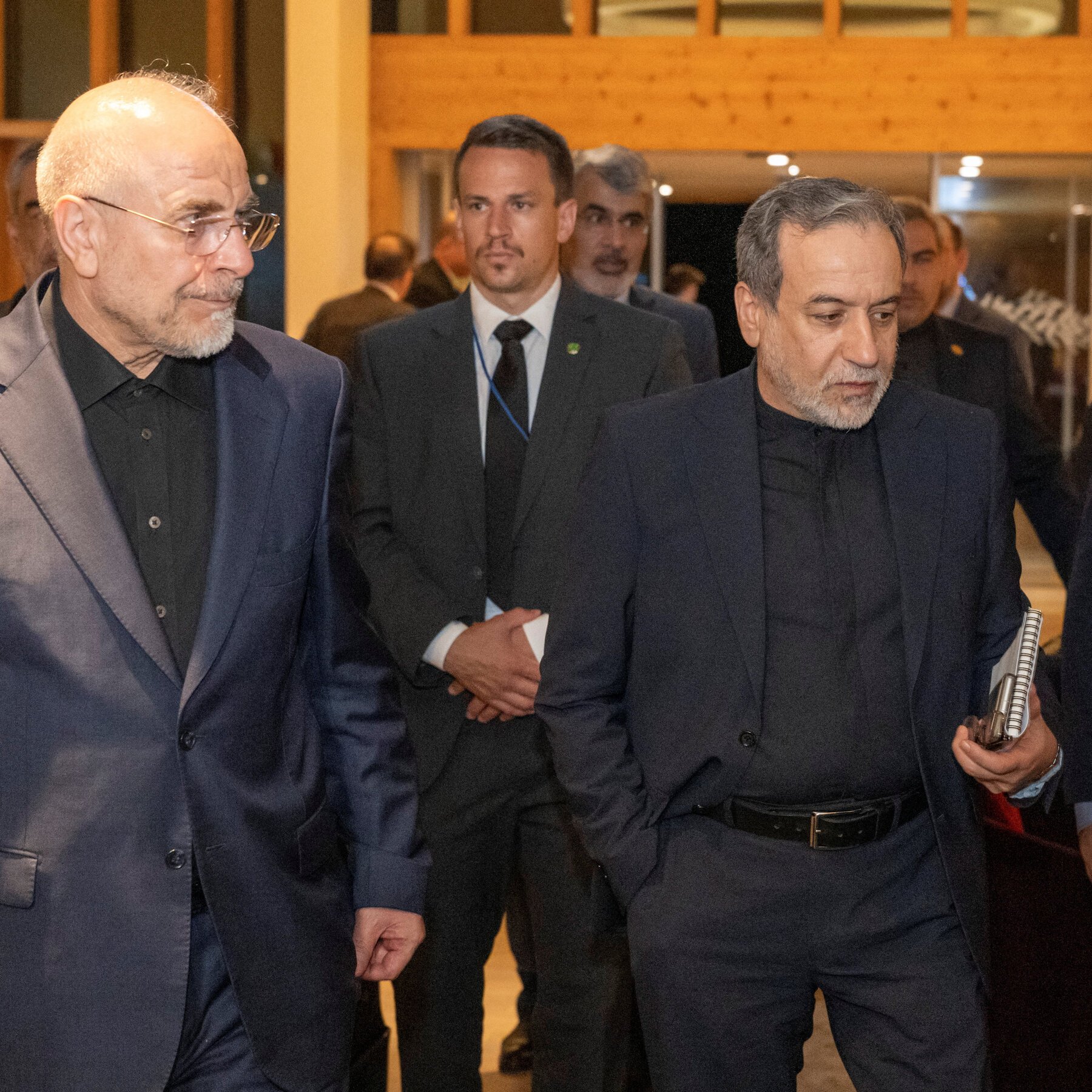 Two older men in dark suits walk side by side through an indoor venue, accompanied by several suited aides in the background.