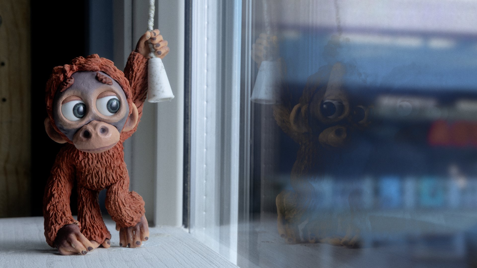 A small brown monkey figurine with large eyes hangs from a white pull-cord beside a window, its reflection visible in the glass.