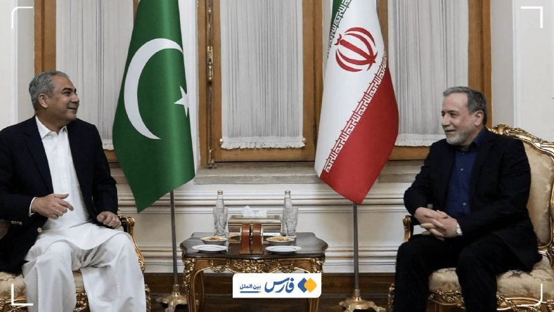 Two men sit facing each other in ornate chairs, with the green Pakistani flag and Iran's red, white, and green flag displayed behind them in a formal meeting room.