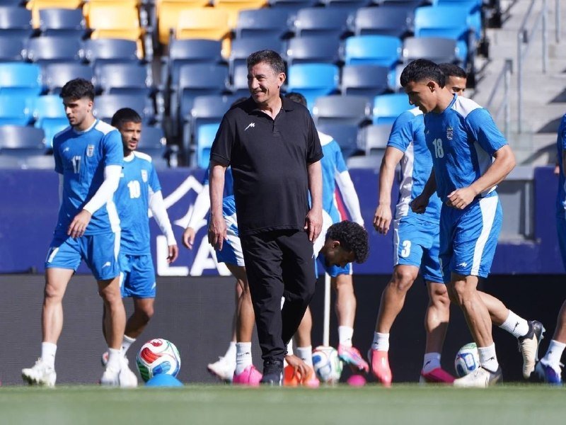 A smiling man in black walks among soccer players in blue uniforms on a grass field with empty stadium seats behind them.