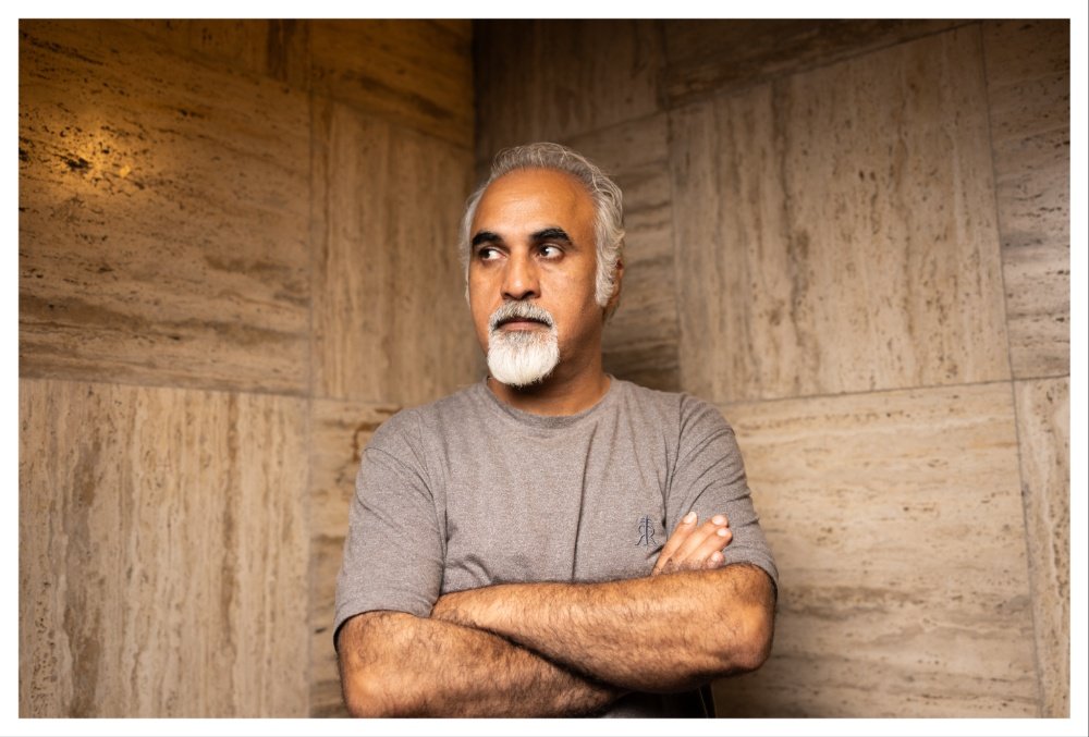 A gray-haired, bearded man in a gray t-shirt stands with arms crossed against a tan stone-tiled wall, looking to his right.