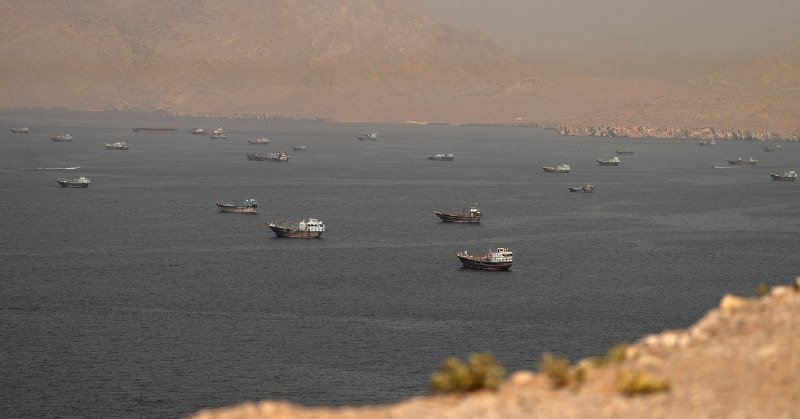 Dozens of cargo ships and tankers are anchored in hazy waters off a rugged, arid coastline.