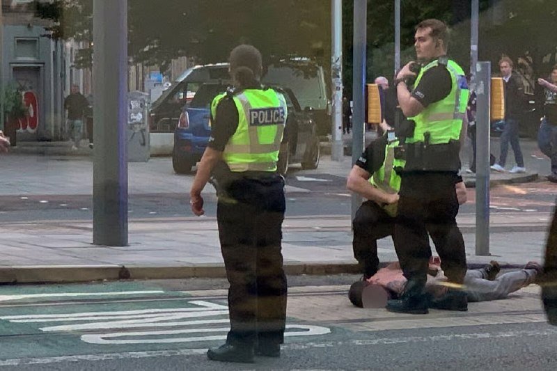 Police officers in high-visibility vests restrain a person face-down on a city street near parked vehicles and a crosswalk.