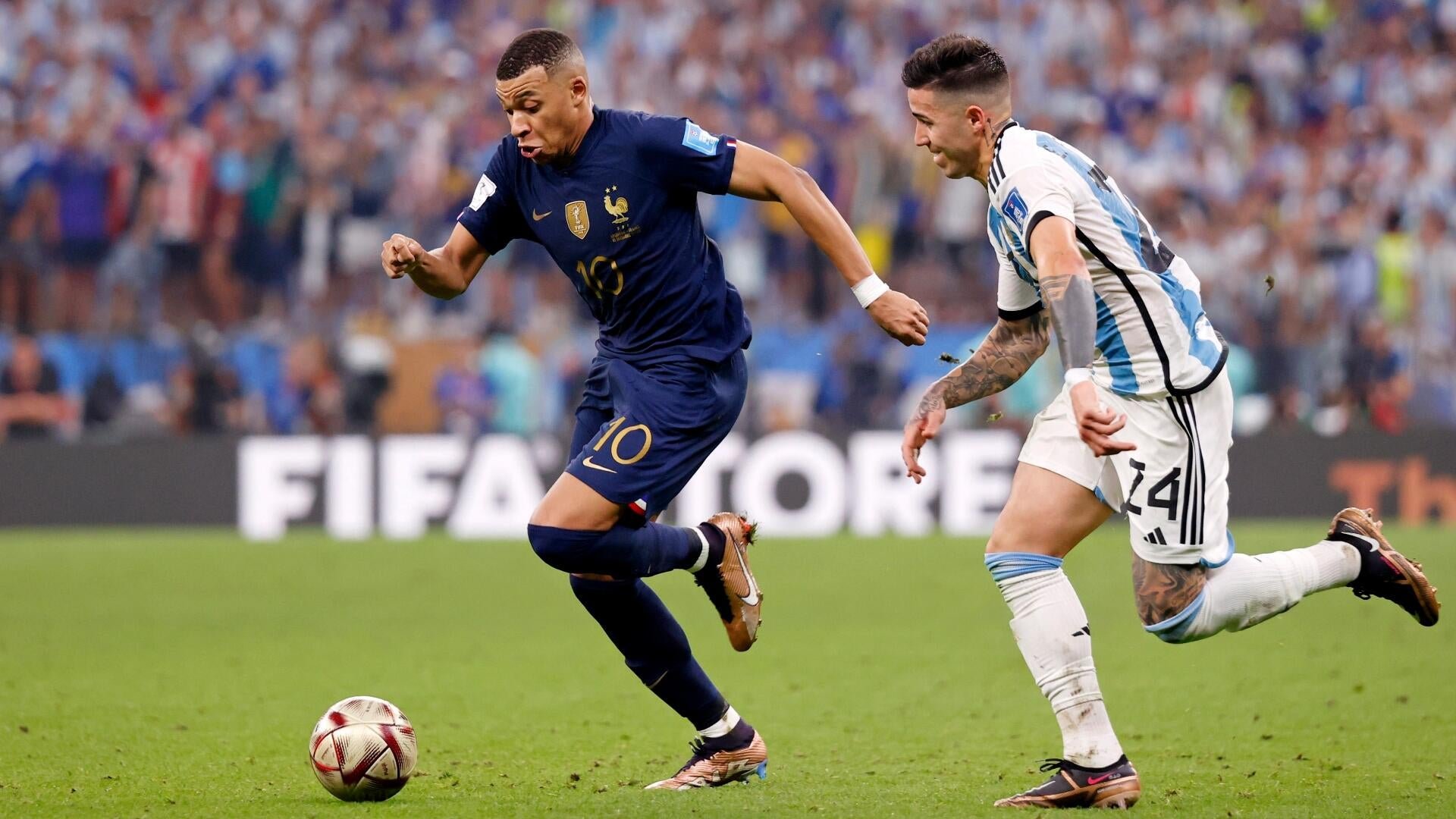 Two soccer players compete on a grass field, one in a dark blue jersey numbered 10 dribbling a ball while another in a white and light blue striped jersey numbered 24 pursues him.
