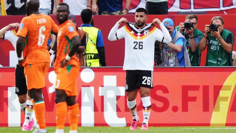A soccer player wearing a white jersey with number 26 pulls the collar of his shirt toward his face, while players in orange jerseys walk past and photographers take pictures behind a barrier.