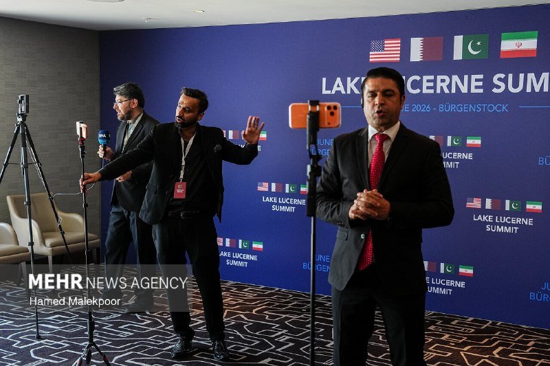 Three men in suits stand before a blue backdrop reading "LAKE LUCERNE SUMMIT" with flags, as one gestures and another speaks with clasped hands.