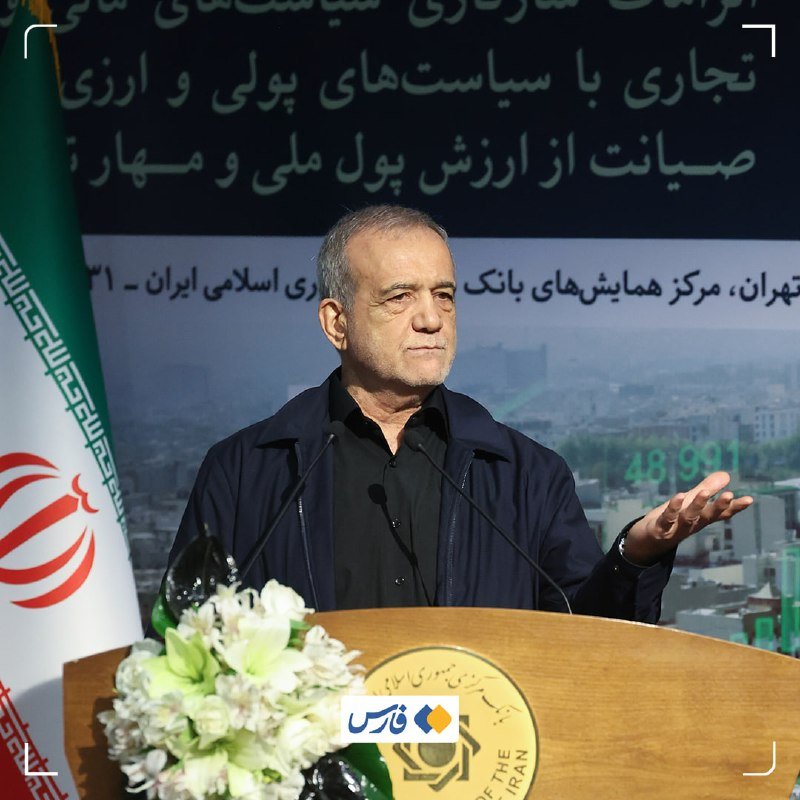 A gray-haired man in a black shirt speaks at a podium with microphones, gesturing with one hand, in front of a Persian banner and the Iranian flag.