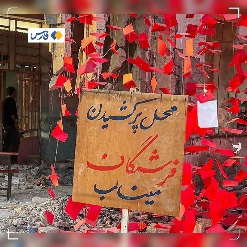 A yellow sign with Persian text stands amid rubble and red paper scraps hanging from strings, with a person visible near a doorway in the background.