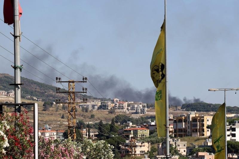 Dark smoke rises above a hillside town, viewed past yellow flags, power lines, utility poles, and flowering plants in the foreground.