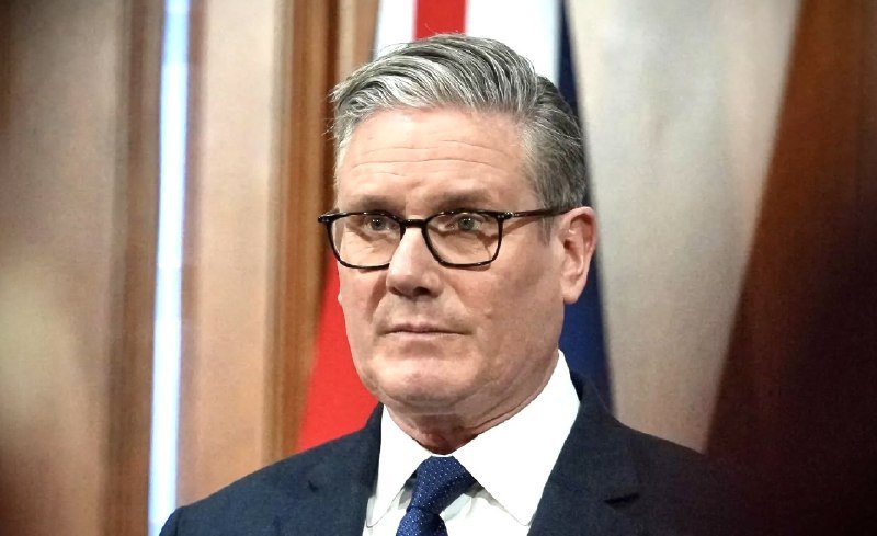 A man with grey hair and glasses, wearing a dark suit and blue tie, stands in front of a partially visible flag.