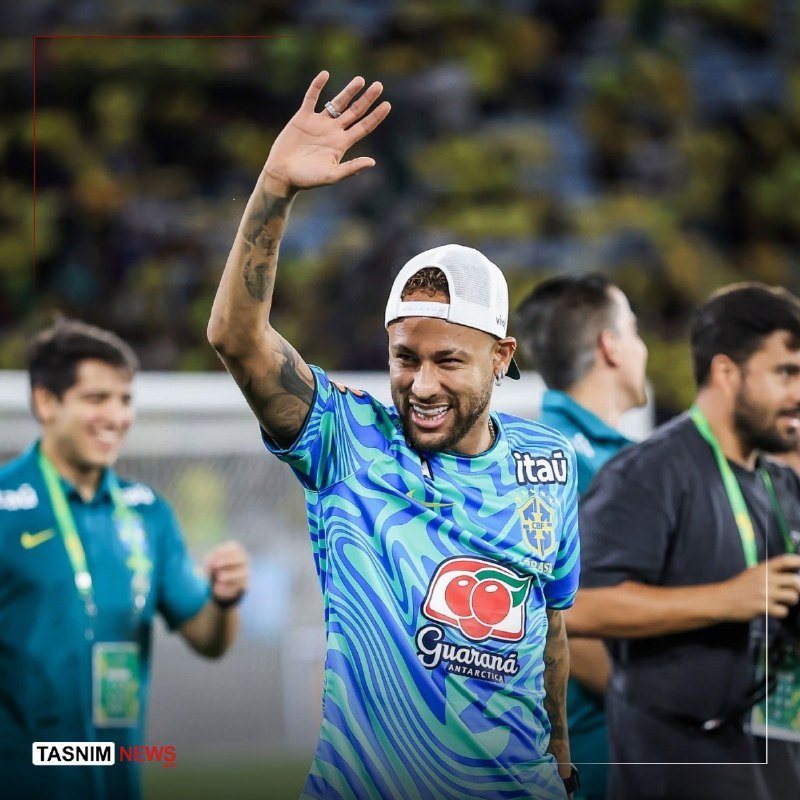 A smiling soccer player wearing a blue Brazilian national team training jersey and white backwards cap waves his tattooed arm toward the crowd.