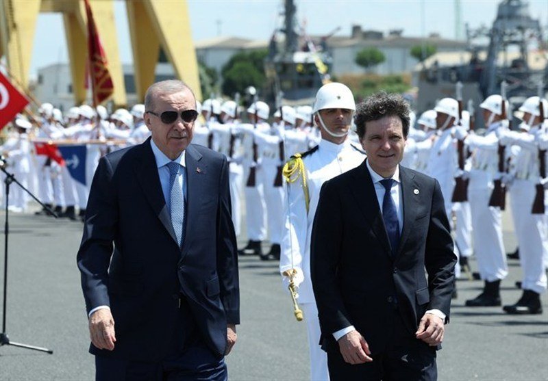 Two men in dark suits walk side-by-side past rows of white-uniformed naval sailors standing at attention, with warships and yellow cranes visible in the background.