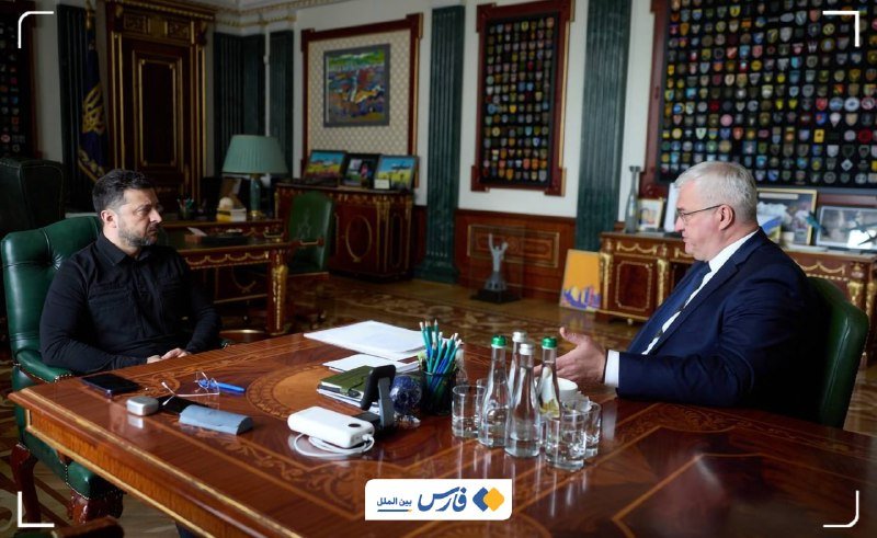 Two men sit across a wooden table in an ornate office with green leather chairs, conversing amid documents, water bottles, and pencils, with framed displays on the walls.