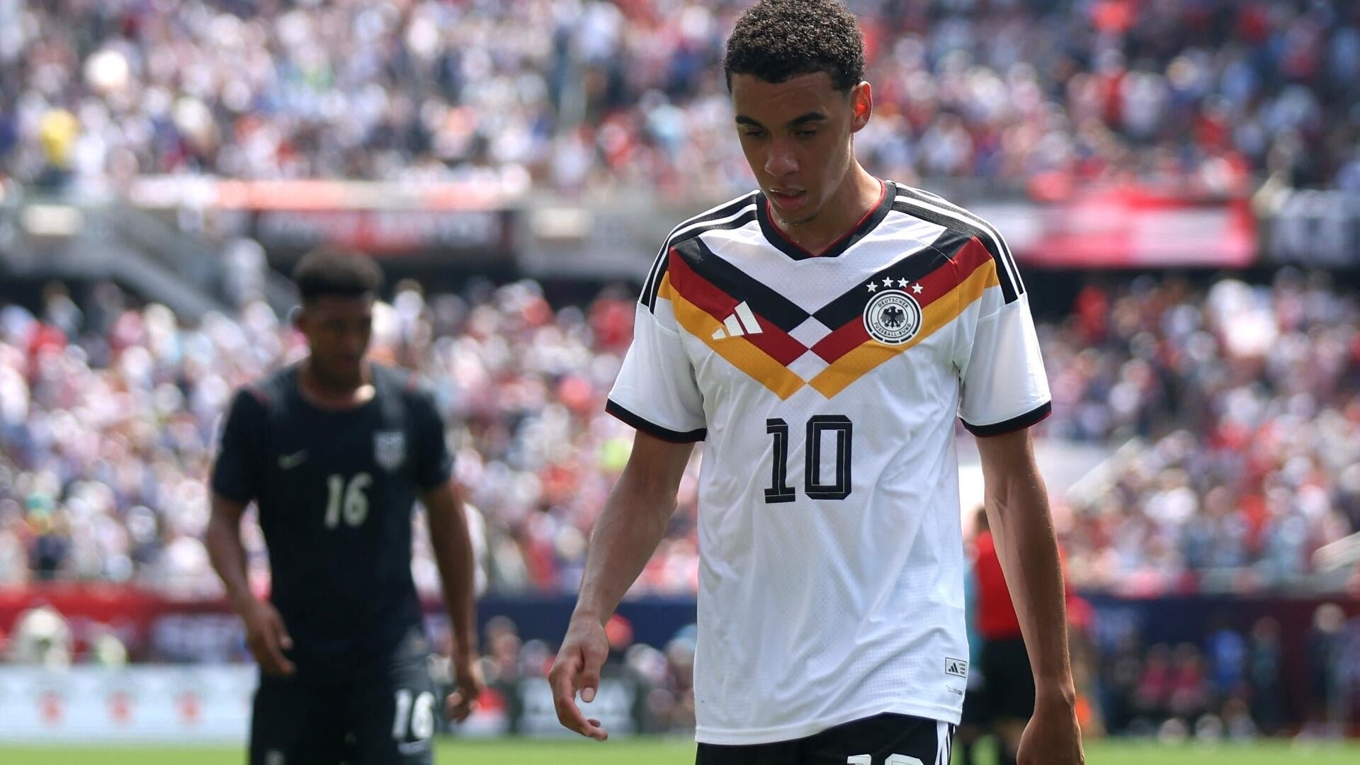 A soccer player wearing a white Germany national team jersey with the number 10 walks on the pitch, while an opposing player in a dark number 16 jersey stands nearby before a stadium crowd.