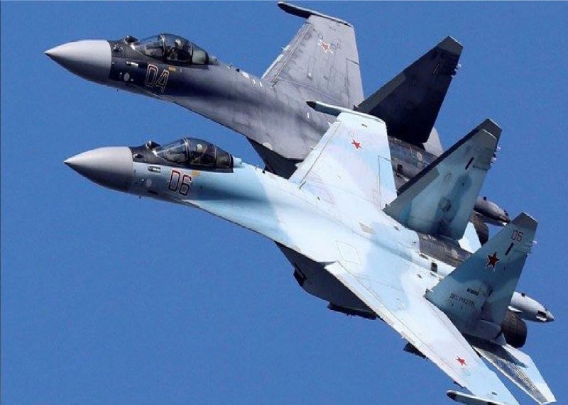 Two military fighter jets, one gray marked "04" and one light blue marked "06," fly in close formation against a clear blue sky.