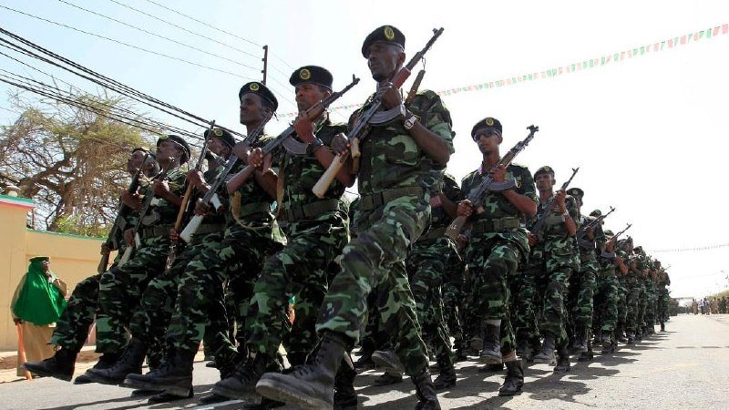 Soldiers in camouflage uniforms and berets march in formation down a street, carrying rifles, with overhead wires and festive bunting visible in the background.