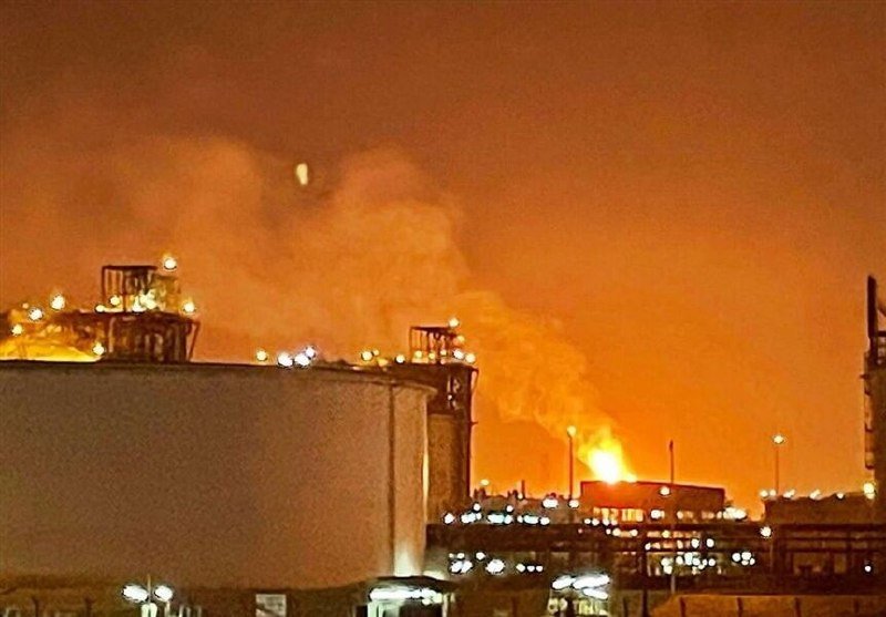 An industrial facility is engulfed in flames and thick smoke at night, with bright orange light illuminating the sky above large storage tanks and surrounding structures.