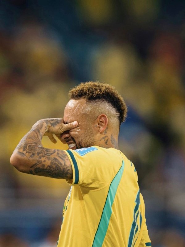 A tattooed soccer player in a yellow and green jersey covers his eyes with his hand on a stadium field.