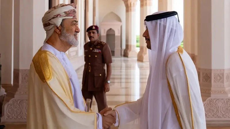 Two men in traditional Arab attire shake hands and smile in an arched corridor, while a uniformed officer stands at attention in the background.