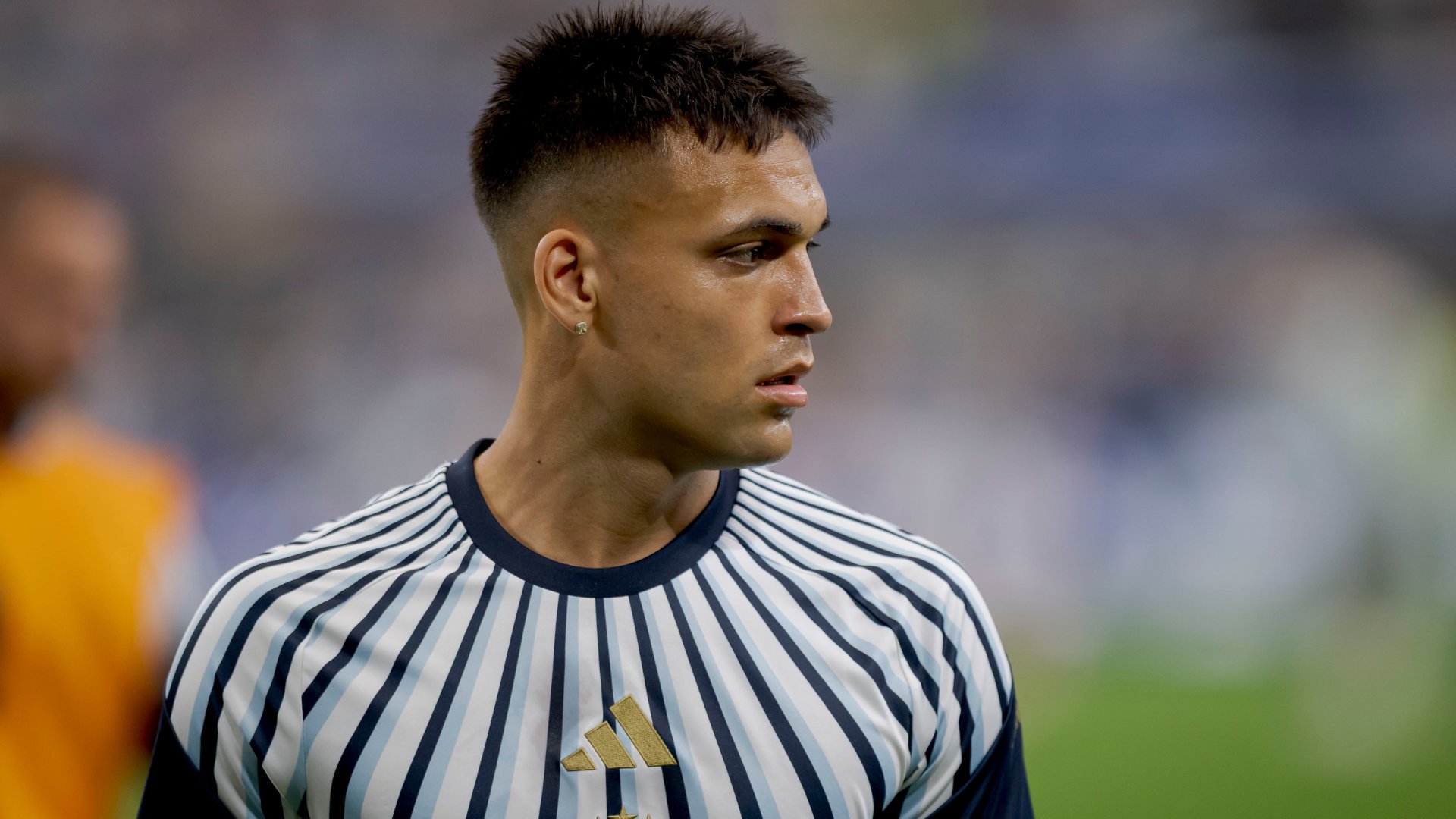 A player in a black-and-white striped jersey with a gold logo stands on a field, looking to the side, with a blurred figure in yellow behind him.