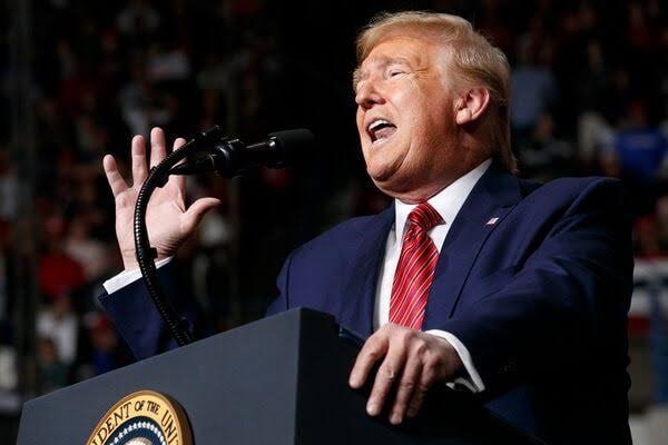 A man in a dark suit and red tie speaks at a podium bearing a presidential seal, gesturing with his right hand before a blurred crowd.