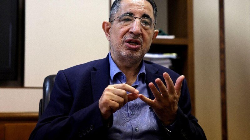 A bespectacled man in a dark blazer and blue shirt gestures with both hands while speaking during an indoor interview.