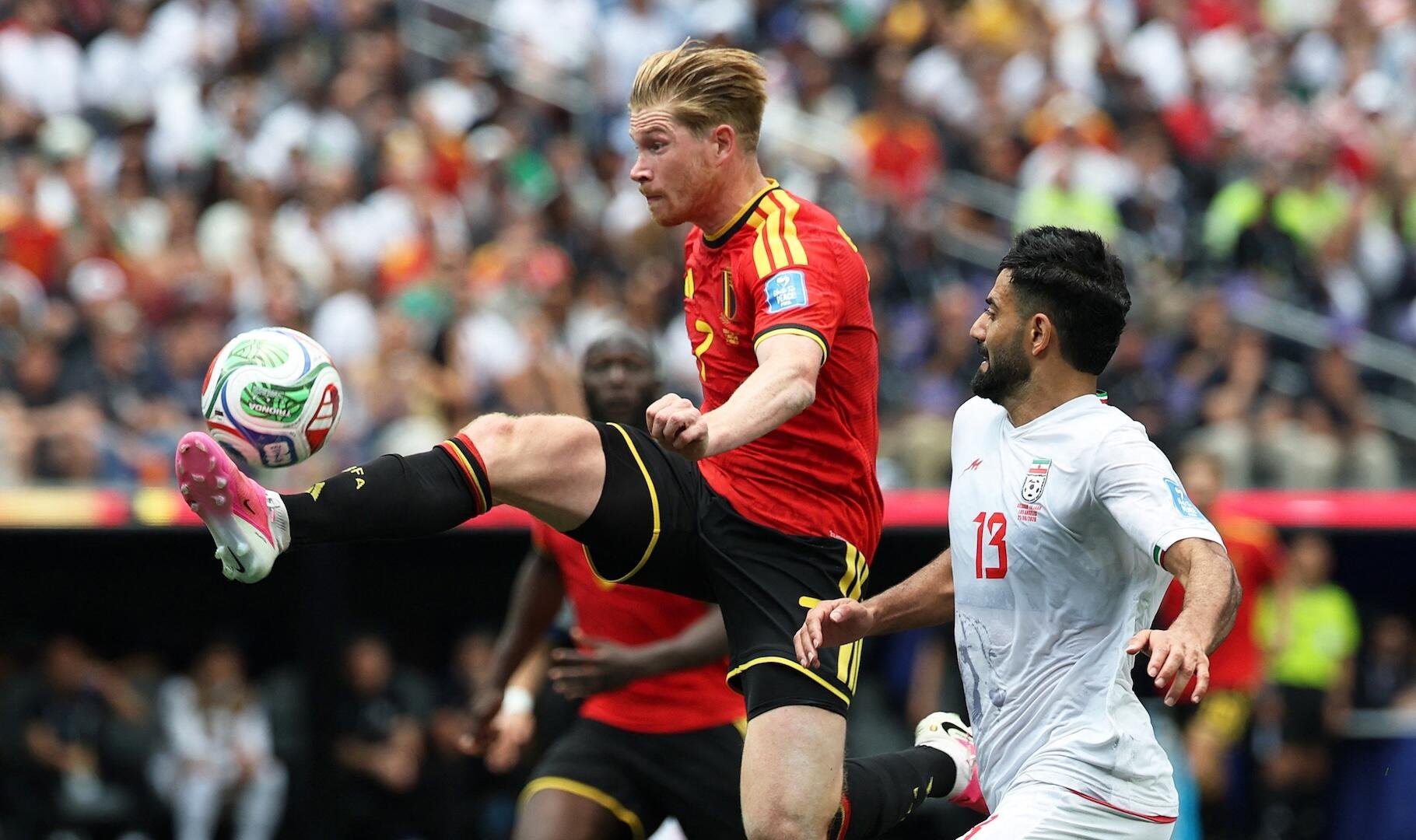 A soccer player in a red and black jersey kicks the ball mid-air while an opponent in a white jersey defends, during a match in a crowded stadium.