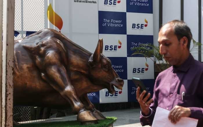 A bronze bull statue stands beside a man in a purple shirt looking at his phone, in front of a backdrop displaying the "BSE" logo and "The Power of Vibrance" text.