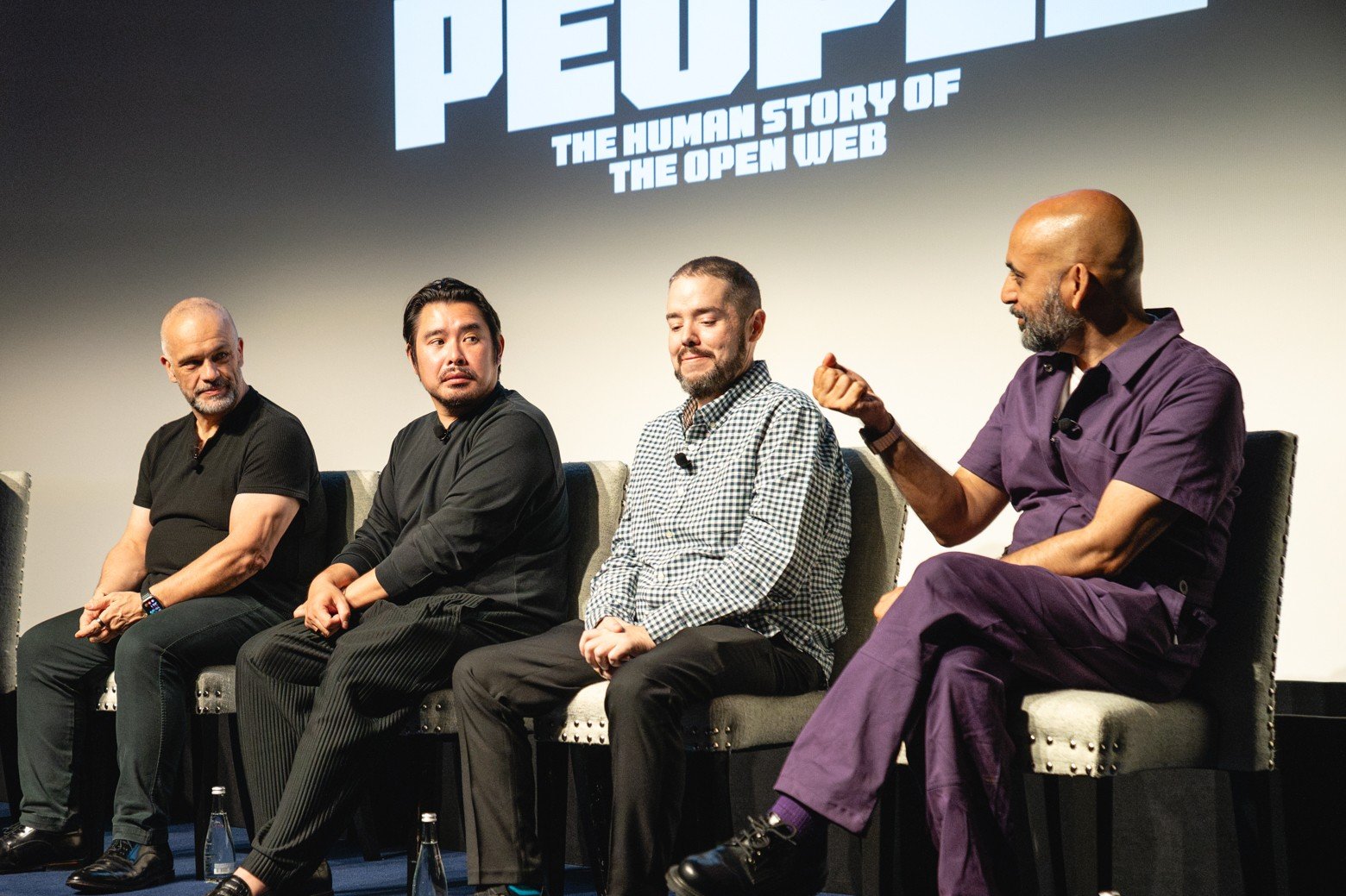Four men sit on gray chairs on a stage during a panel, with one gesturing while speaking; a large screen behind them displays "PEOPLE: THE HUMAN STORY OF THE OPEN WEB."