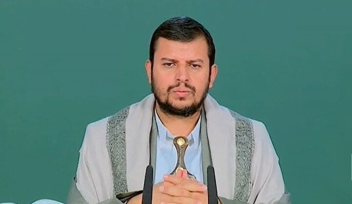 A bearded man in a light blue shirt and white shawl speaks at two microphones while holding a small object against a green backdrop.
