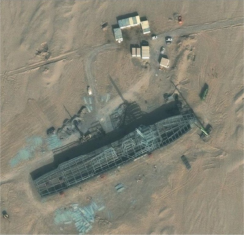 Aerial view of a construction site in a desert landscape, showing a long metal framework structure, nearby small buildings, vehicles, and dirt roads.