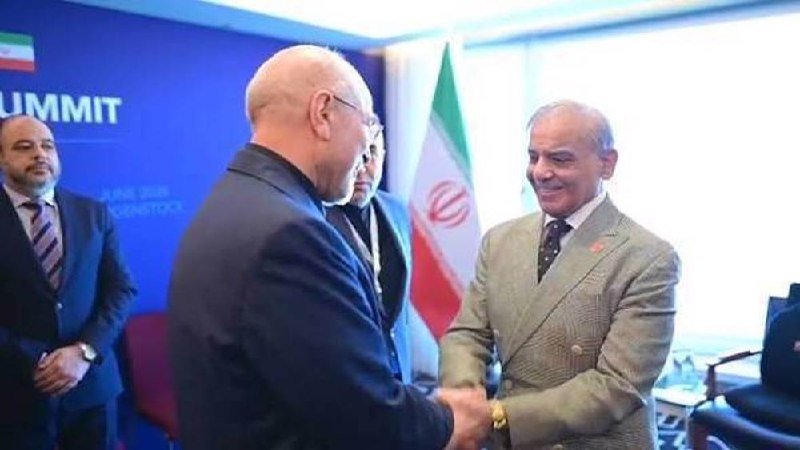 Two men in suits shake hands and smile in front of an Iranian flag and a blue banner reading "SUMMIT," with two other men standing nearby in an indoor setting.