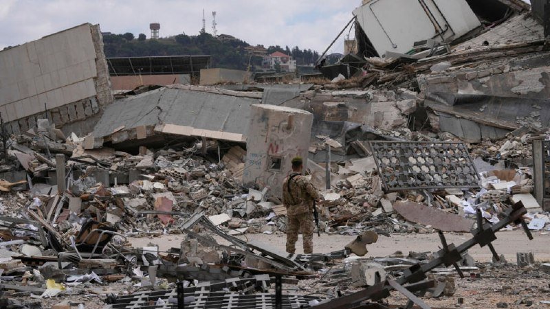 A soldier in camouflage stands amid the extensive rubble of collapsed concrete buildings and scattered debris under a partly cloudy sky.