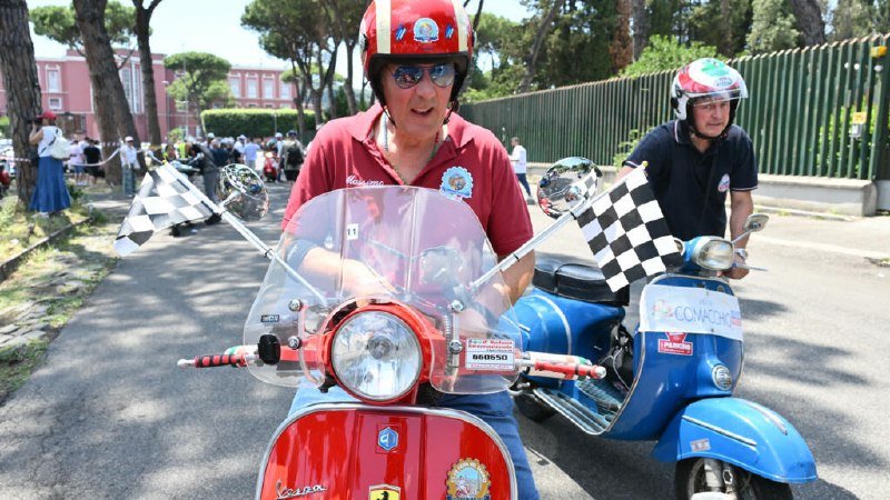 Two helmeted men ride red and blue Vespa scooters down a sunlit street, both displaying checkered racing flags.