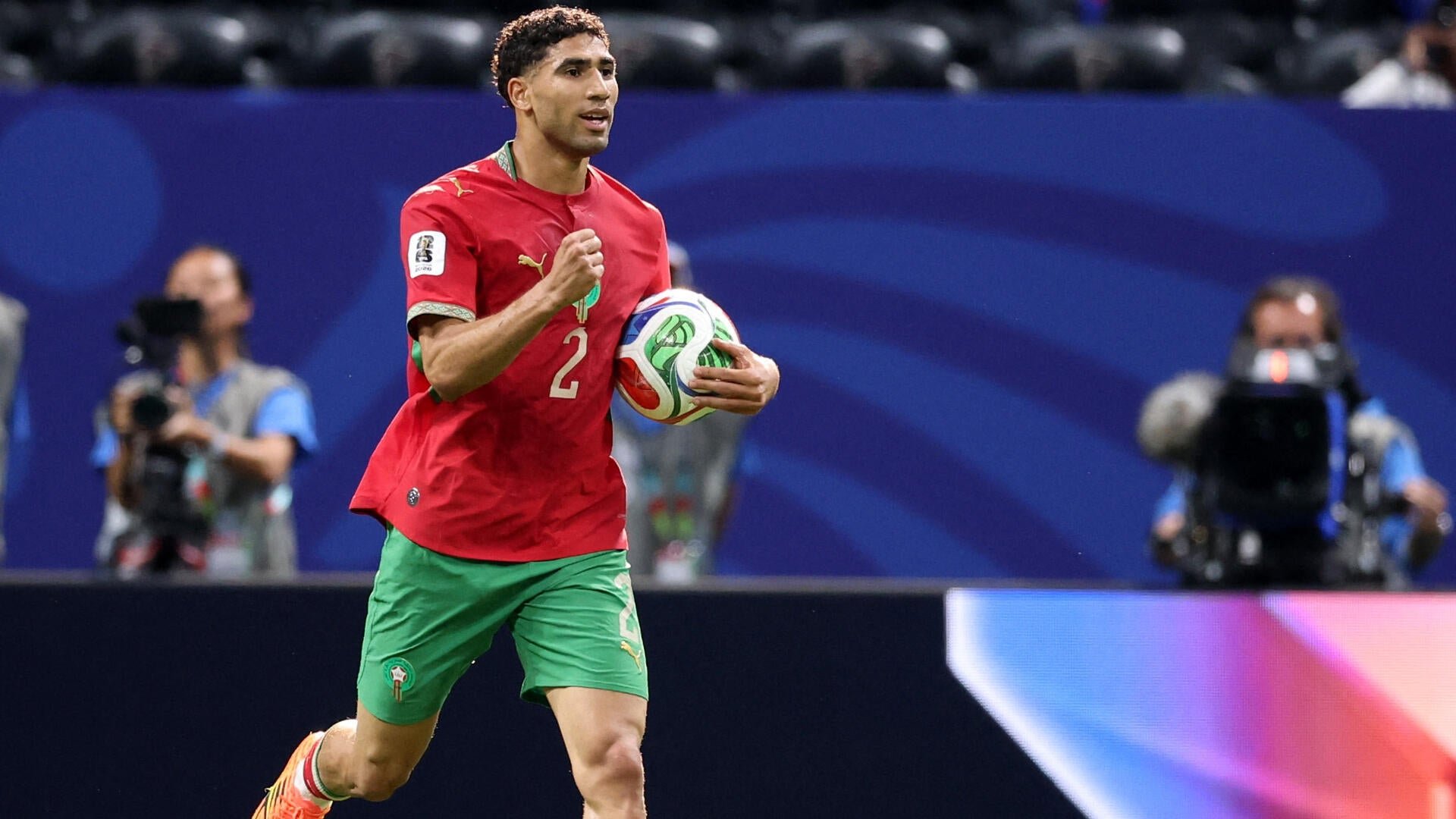A soccer player in a red jersey numbered 2 and green shorts runs across a pitch clutching a soccer ball, with cameramen visible in the background.