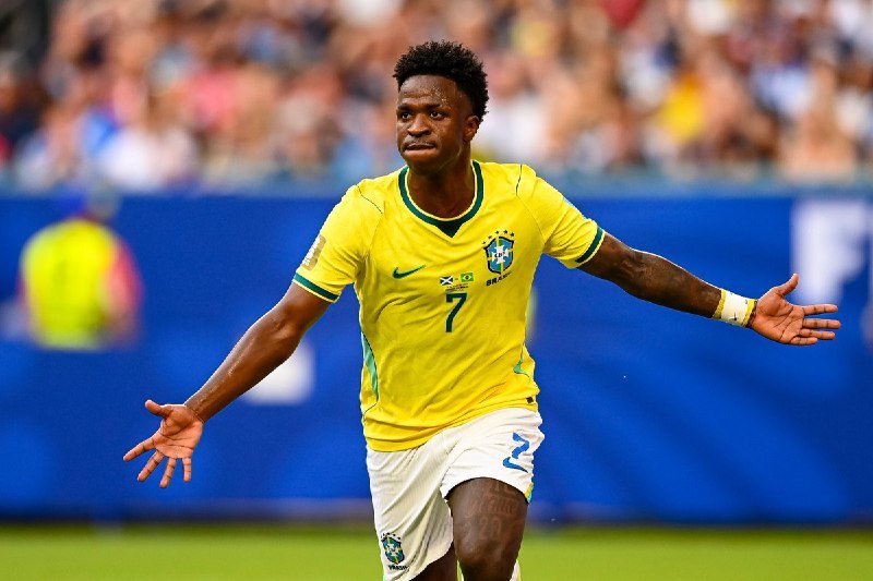 A soccer player in a yellow Brazil jersey with the number 7 celebrates with outstretched arms on the pitch before a blurred crowd.